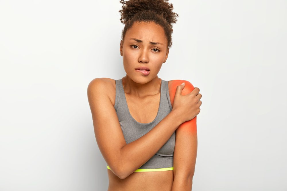 Displeased curly haired woman bites lips, touches painful shoulder, suffers from injury during going in for sport in gym, wears grey top, isolated on white background. Health problems, medical concept