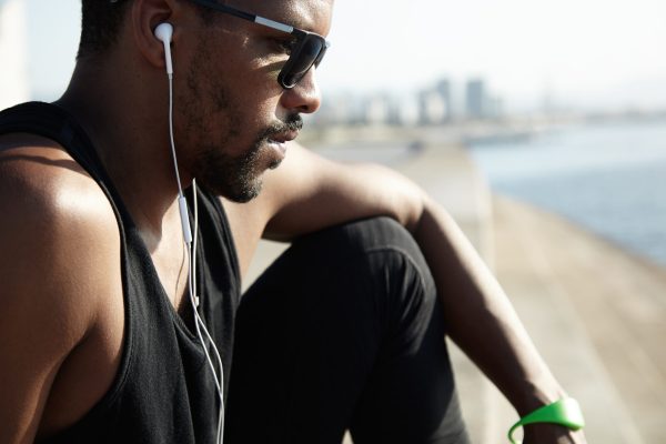 Profile portrait of young black runner in earphones relaxing after his morning workout in open air, listening to music, looking tired and exhausted, sitting on pavement, breathing fresh sea air